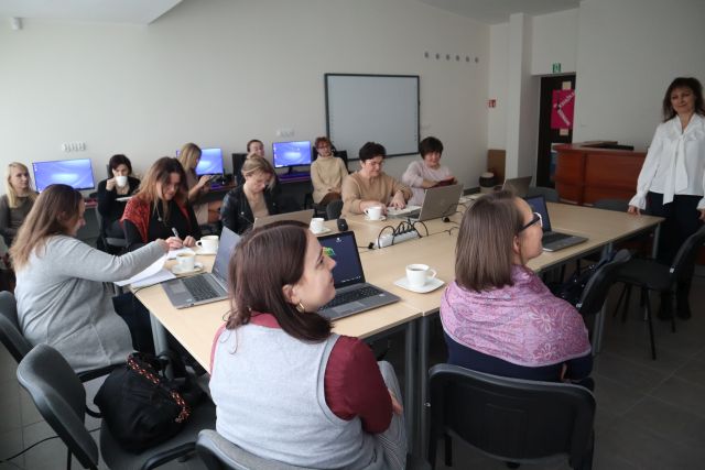 uczestnicy szkolenia siedzą przy brązowych stołach, przed nimi na stołąch stoją otwarte laptopy