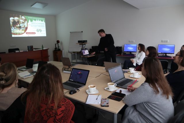 uczestnicy szkolenia siedzą przy brązowych stołąch, przed sobą mają laptopy, w tle stoi mężczyzna ubrany na czarno