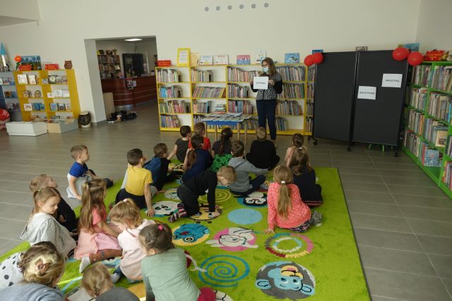 na żółtym dywanie siedzą tyłem dzieci różnie ubrane, przed nimi stoi kobieta w ciemnym stroju, za nią żółte regały z książkami