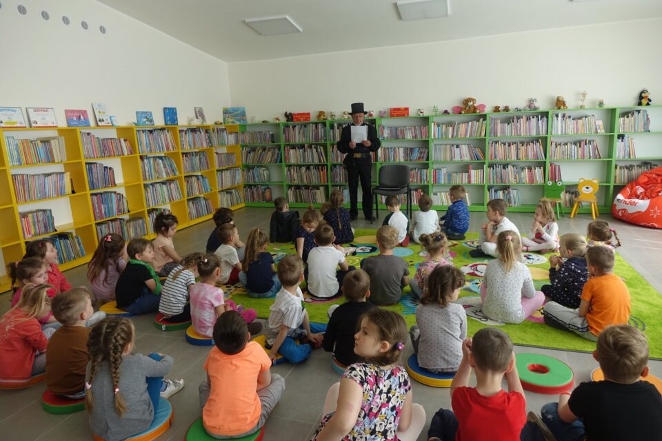 na podłodze i na zielonym dywanie tyłem siedzą dzieci, przed nimi stoi na czarno ubrany kominiarz, któey w dłoniach trzyma kartkę, za nim od lewej strony żółte regały i zielone regały