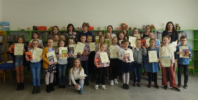 dzieci i młodzież stoją w kilku rzędach i trzymają w dłoniach dyplomy i książki, za n imi stoi 6 osób dorosłych, za nimi zielone regały