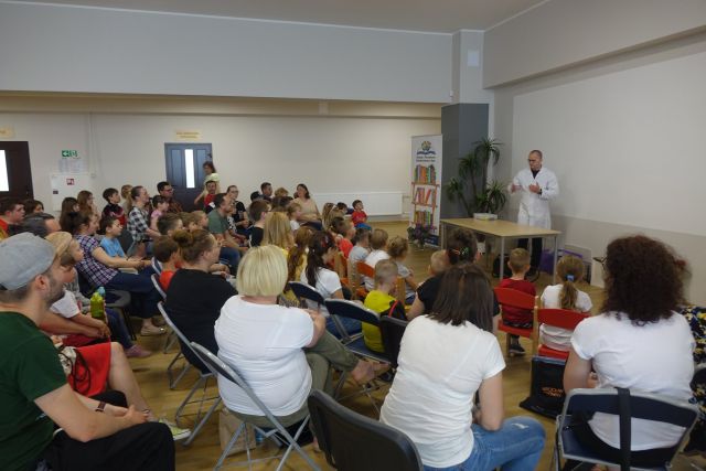 na niebieskich krzesłąch tyłem siedzą dzieci i dorośli, przed nimi w białym fartuchu stoi przed brązowym stołem mężczyzna