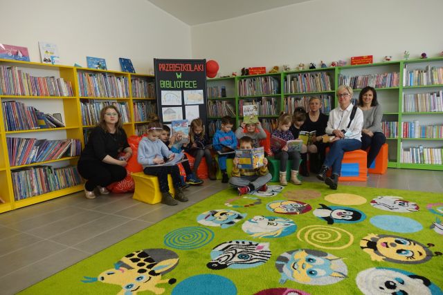 za żółtym dywanem siedzą na kolorowych pufach dzieci i trzy kobiety dorosłe wszyscy różnokolorowo ubrani, za nimi żółte i zielone regały z książkami