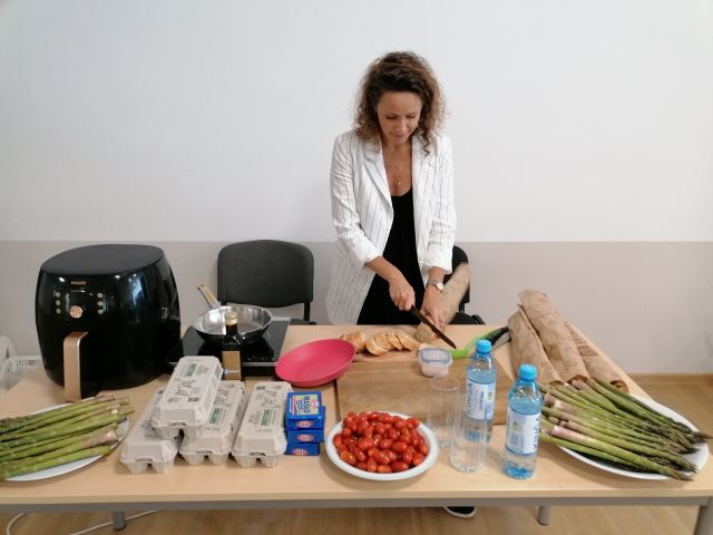 za plecami stojącej kobiety jasna ściana, kobieta ubrana w białą marynarkę i czarną sukienkę, stoi przed brązowym stołem, na którym leżą artykuły spożywcze: masło jajka w kartonach, szparagi zielone. Kobieta kroi bagietki. Na stole stoi czarny garnek do przyrządzania potraw