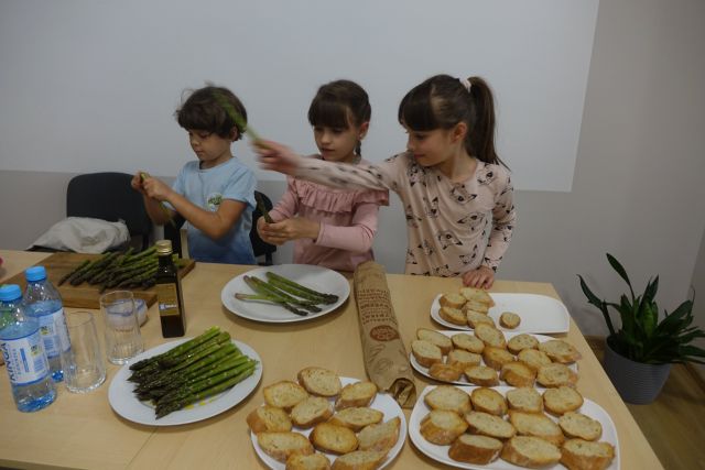 przy brązowym stole stoją dwie dziewczynki i jeden chłopiec 9. letni. Dzieci kroją pomidory i szparagi