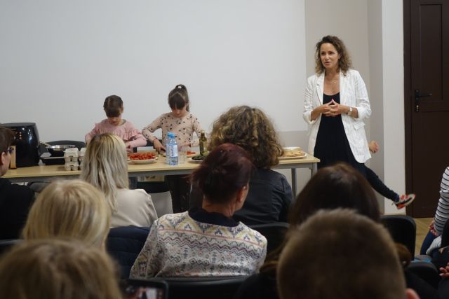 przy brązowym stole stoją dwie dziewczynki i kroją pomidory, po prawej stronie stołu stoi kobieta, przed nią tyłem siedzą uczestnicy spotkania