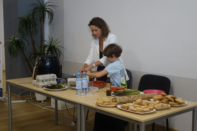 przy brązowym stole stoi kobieta i chłopiec, szykują potrawę - wybijają jajka na jajecznicę, na stole leżą artykuły spożywcze i czarny garnek