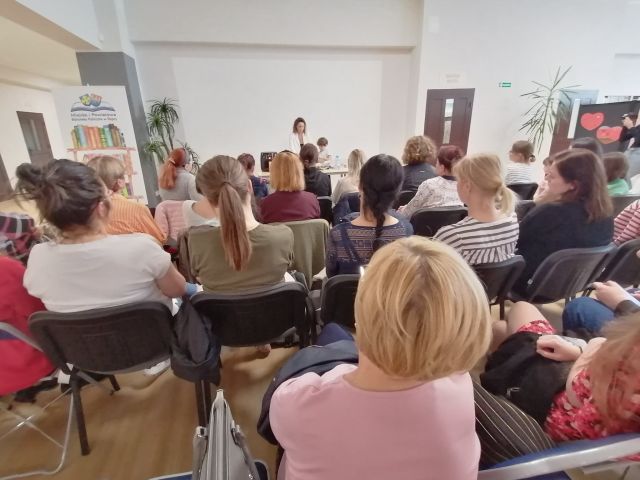 na siwych krzesłąch siedzą tyłem uczestnicy spotkania, przed nimi stoi kobieta i opowiada