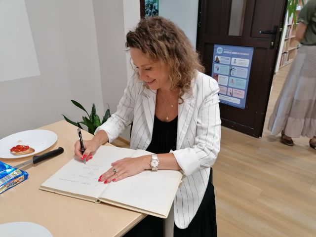 przy brązowym stole na siwym krześle siedzi kobieta w białej marynarce i czarnej sukience i wpisuje się do białej księgi