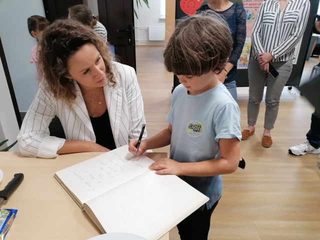 przy stole siedzi kobieta z ręką opartą o stół i patrzy na chłopca w niebieskiej bluzce, który wpisuje się do księgi