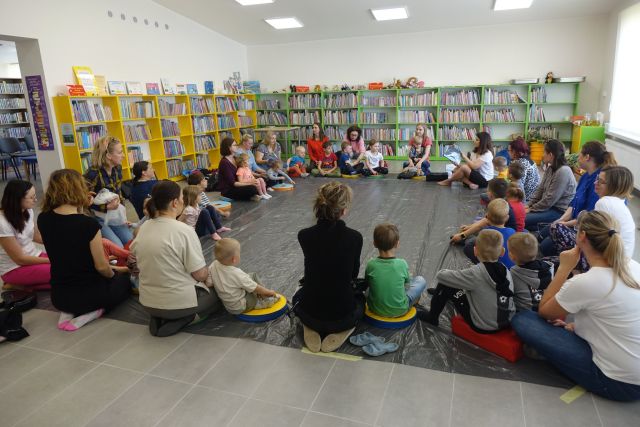 na siwej podłodze siedzą w kole małe dzieci i mamy na kolorowych pufach, za nimi zielone i żółte regały z książkami