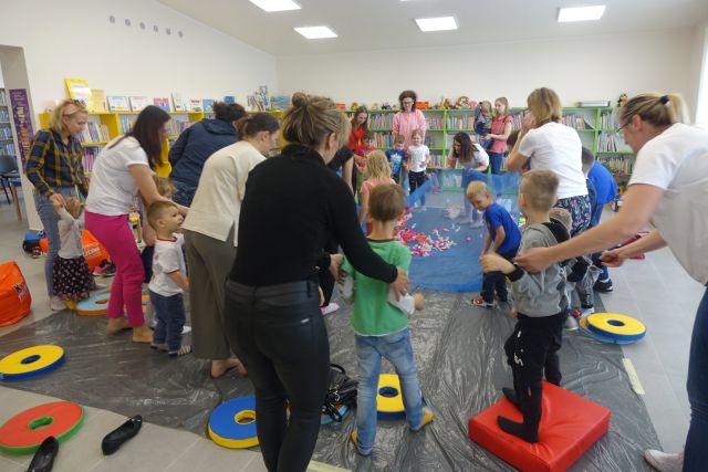 na siwej podłodze zasłoniętej folią stoją dzieci i mamy, w środku niebieski materiał, a na nim kolorowe pióra, za osobami zielone i żółte regały z książkami