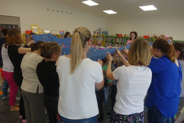 dorosłe osoby stoją w kółku i trzymają w dłoniach niebieski materiał, na nim kolorowe pióra, pod materiałem dzieci biegają