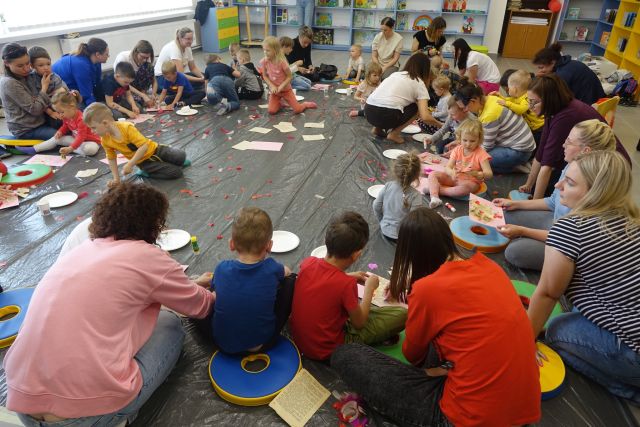 na siwej folii malarskiej siedzą dzieci i ich mamy, w środku różne przedmioty: pióra, kartki, papierowe talerzyki