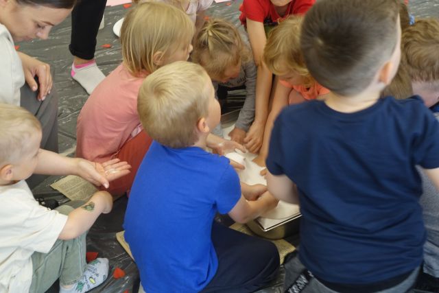 na siwej podłodze kucała dzieci, w środku na podłodze miska z białym cukrem, którym bawią się dzieci