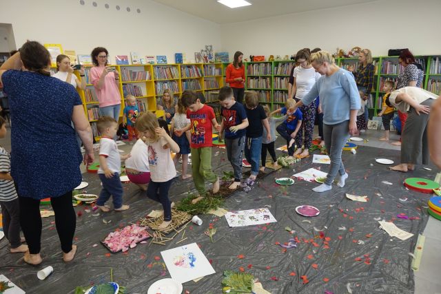 na siwej podłodze ułożona ścieżka z miskami z liśćmi, kwiatami, drewienkami, kaszą, mąką, bzem, po którą bosymi stopami spacerują dzieci i mamy