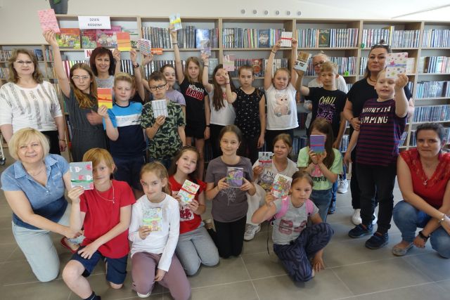 na tle brązowych regałów z książkami stoją dzieci z książkami wykonanymi przez siebie w dłoniach, przed nimi kucają dzici z książkami obrazkowymi wykonanymi przez siebie