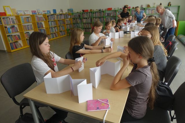 przy brązowych stołach siedzą dzieci, na stołach kartki i przybory do malowania, za nimi zielone i żółte regały