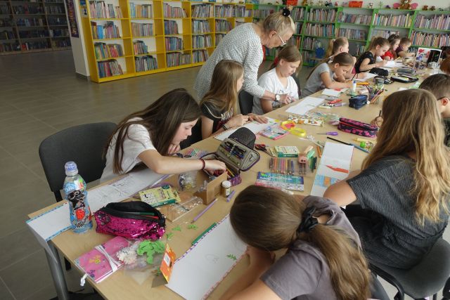 przy brązowych stołach siedzą dzieci, na stołach kartki i przybory do malowania, za nimi zielone i żółte regały