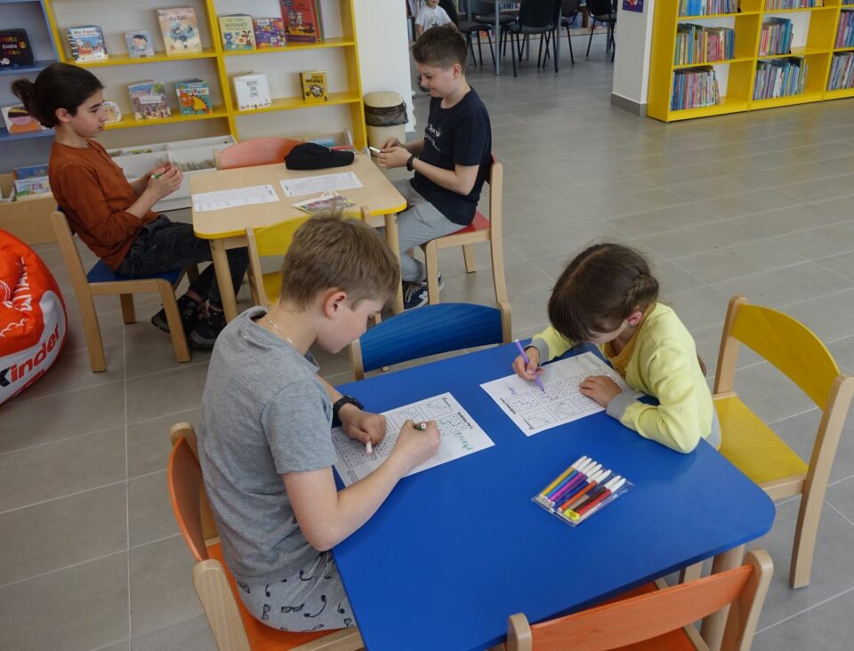 przy niebieskim i brązowym stole siedzą dzieci, na stołach mają kartki po których piszą, za nimi żółte regały