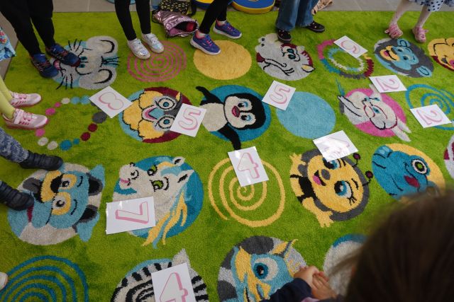 na zielonym dywanie leża ponumerowane białe kartki, w koło widać stópki dzieci