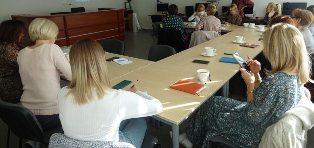 kobietysiedzą przy brązowych stołach i notują w zeszytach, w tle biurka z komputerami