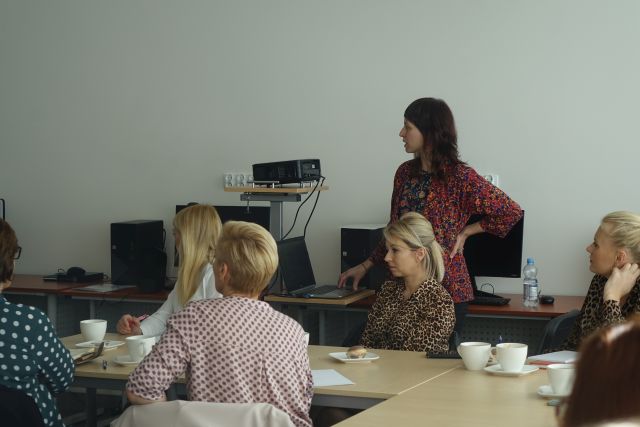 kobiety siedzą przy brązowych stołach, na których stoją filiżanki białe i zeszyty, za nimi stoi kobieta w bluzce czerwonej w kwiaty, w tle biurka z komputerami