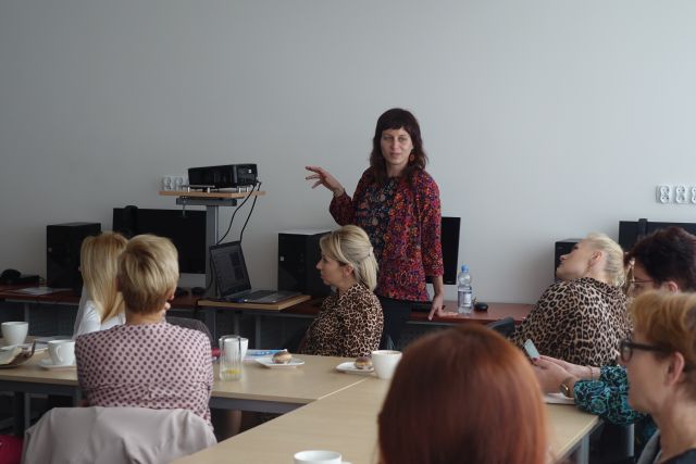 przy brązowych stołach siedzą kobiety, na stołach zeszyty i białę filiżanki, za nimi stoi kobieta w czerwonej bluzce w kwiaty, w tle biurka z komputerami