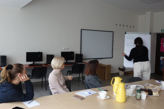 przy brązowych stołach siedzą uczestniczki szkolenia, przed nimi przy tablicy stoi kobieta, w tle biurka z komputerami