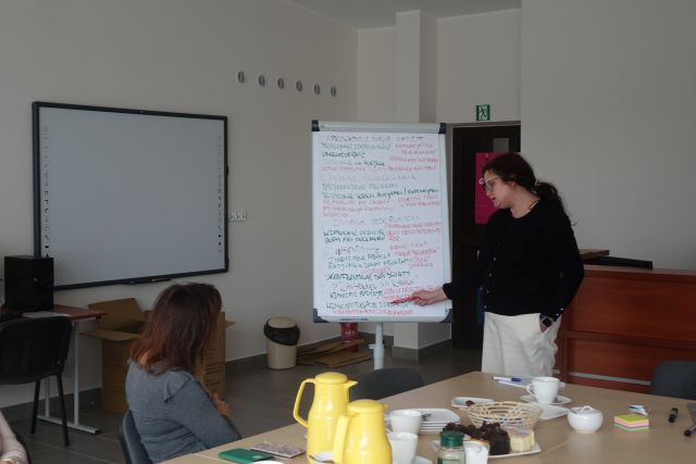 przy brązowych stolikach siedzą uczestniczki szkolenia, przed nimi przy tablicy stoi kobieta w czarnej bluzce i białych spodniach, w tle biurka z komputerami