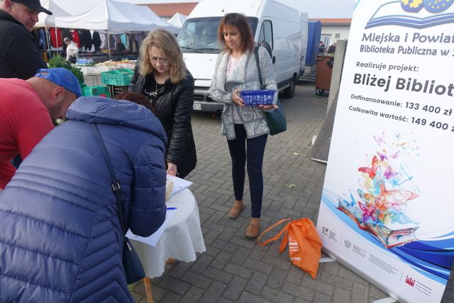 Kobita w granatowej kurtce pochylana nad stolikiem z jasnym obrusem wypełnia ankietę, przed nią stoją dwie kobiety w czarnej kurtce i swetrze w norweskie wzory, z prawej strony baner z nazwą projektu, w tle duży biały samochód bus