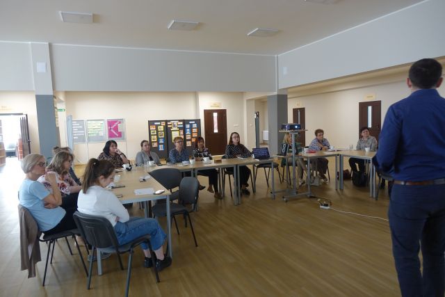 kobiety siedzą przy stołach, przed nimi stoi mężczyzna w niebieskiej koszuli