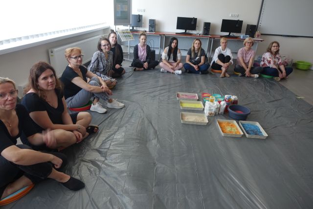 Uczestnicy szkolenia siedzą na siwej podłodze, na środku pojemniki z różnymi sypkimi produktami, w tle biurka z komputerami