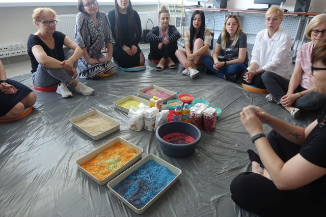 Uczestnicy szkolenia siedzą na siwej podłodze, na środku pojemniki z różnymi sypkimi produktami, w tle biurka z komputerami