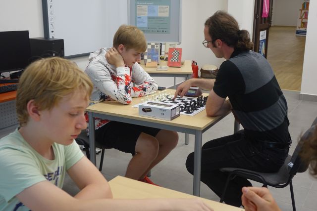 przy brązowych stołach siedzą szachiści, na stołach szachy i zegary