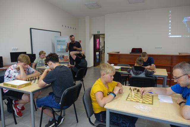 przy brązowych stołąch siedzą szachiści, na stołach szachy, w tle stoi mężczyzna, po prawej stronie w tle brązowe biurko