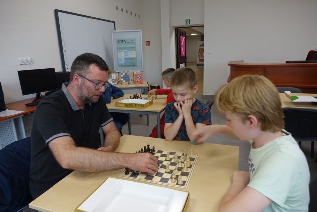 przy stołąch siedzą dzieci i trener szachowy, grają w szach, na stołąch szacy