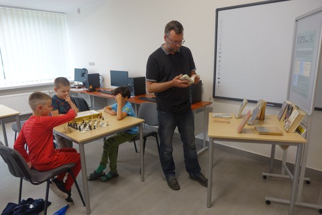 przy stołąch siedzą i grają mali szachiści, przy stole z książkami o szachach stoi mężczyzna i przegląda książkę