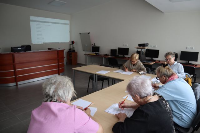 przy brązowych stołach siedzą starsze kobiety i piszą na kartkach, przed nimi na ścianie ekran z prezentacją i brązowe biurko