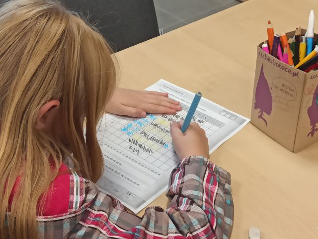 przy brązowym stole tyłęm siedzi dziewczynka w jasnych włosach i pisze na kartce