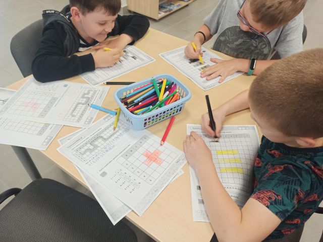 na brżowym stole leżą katki z krzyżówkami, dzieci wypełniają krzyżówki, na środku stołu leży pojemnik nibieski z kredkami