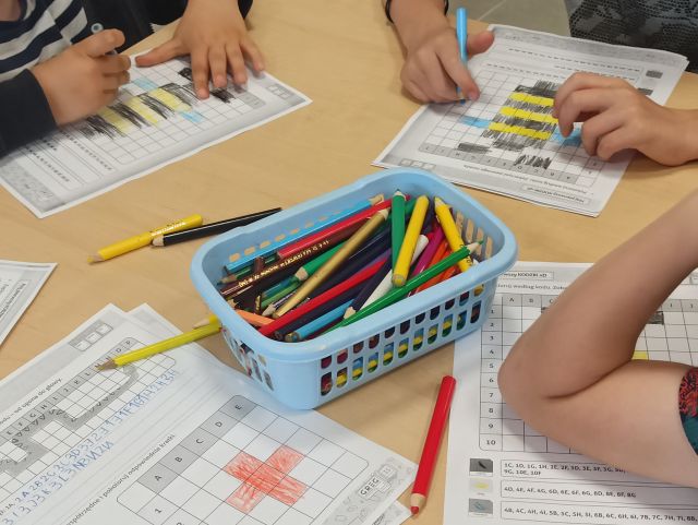 na brązowym stole leżą kartki z krzyżówkami, dzieci wypełniają je, na śrdoku stołu leży niebieski pojemnik z kredkami