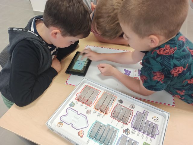 przy stole brązowym siedzą dzieci i patrzą w tablet, na stole leżą kartki z zadaniami