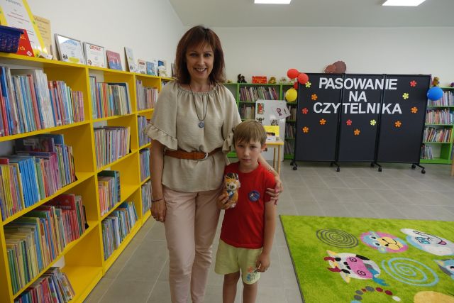 kobieta i chłopiec trzymający w dłoniach sówkę maskotkę. Za nimi żółte regały z książkami, z prawej strony zielony dywan