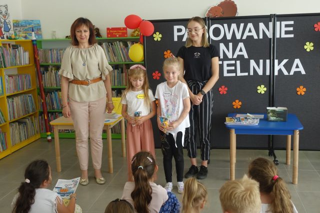 dwie kobiety, pomiędzy nimi dwie dziewczynki trzymające w dłoniach maskotki sówki, za nimi czarna tablica i żółte regały z książkami