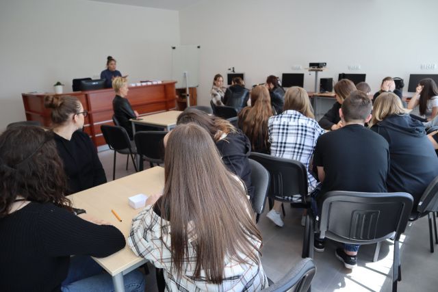 cała sala młodzieży siedzi przy brązowych stołach, za brązowym biurkiem siedzi kobieta