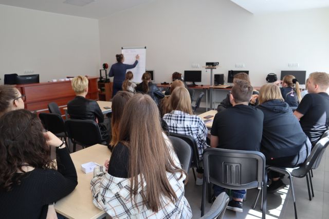 przy brązowych stołach siedzi młodzież , przed nimi przy tablicy stoi kobieta i pisze