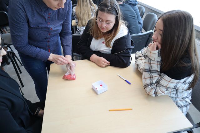przy brązowym stole siedzą trzy młode dziewczyny, przy nich stoi kobieta, na stole kartki i długopisy