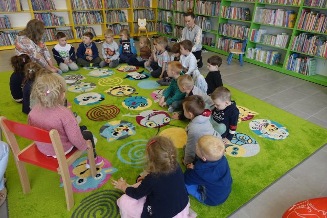 na zielonym dywanie siedzą dzieci i dwie kobiety, za nimi żółte i zielone regały z książkami