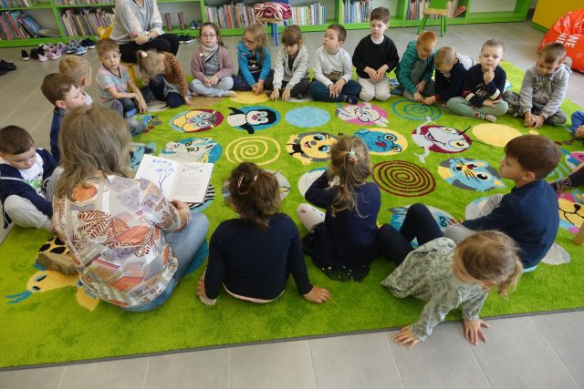 na zielonym dywanie siedzą dzieci i kobieta która czyta książkę, za nimi zielone i żółte regały z książkami